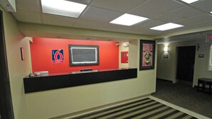 A hotel reception area with a red wall, a reception desk, a framed picture, and a fire extinguisher. - Studio 6 Corporate Extended Stay Buffalo Amherst NY (Buffalo)