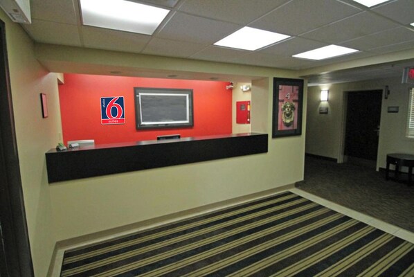 A hotel reception area with a red wall, a reception desk, a framed picture, and a fire extinguisher. - Studio 6 Corporate Extended Stay Buffalo Amherst NY (Buffalo)