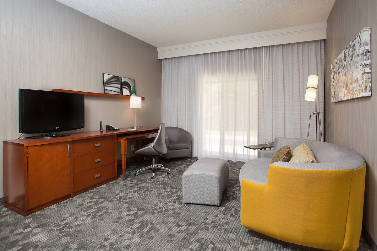 A hotel room with a flat-screen TV, wooden desk, grey armchair, and a yellow sofa.