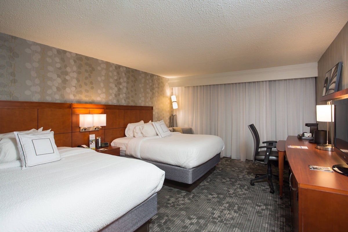 A hotel room with two beds, a desk, a chair, and a large window with curtains.