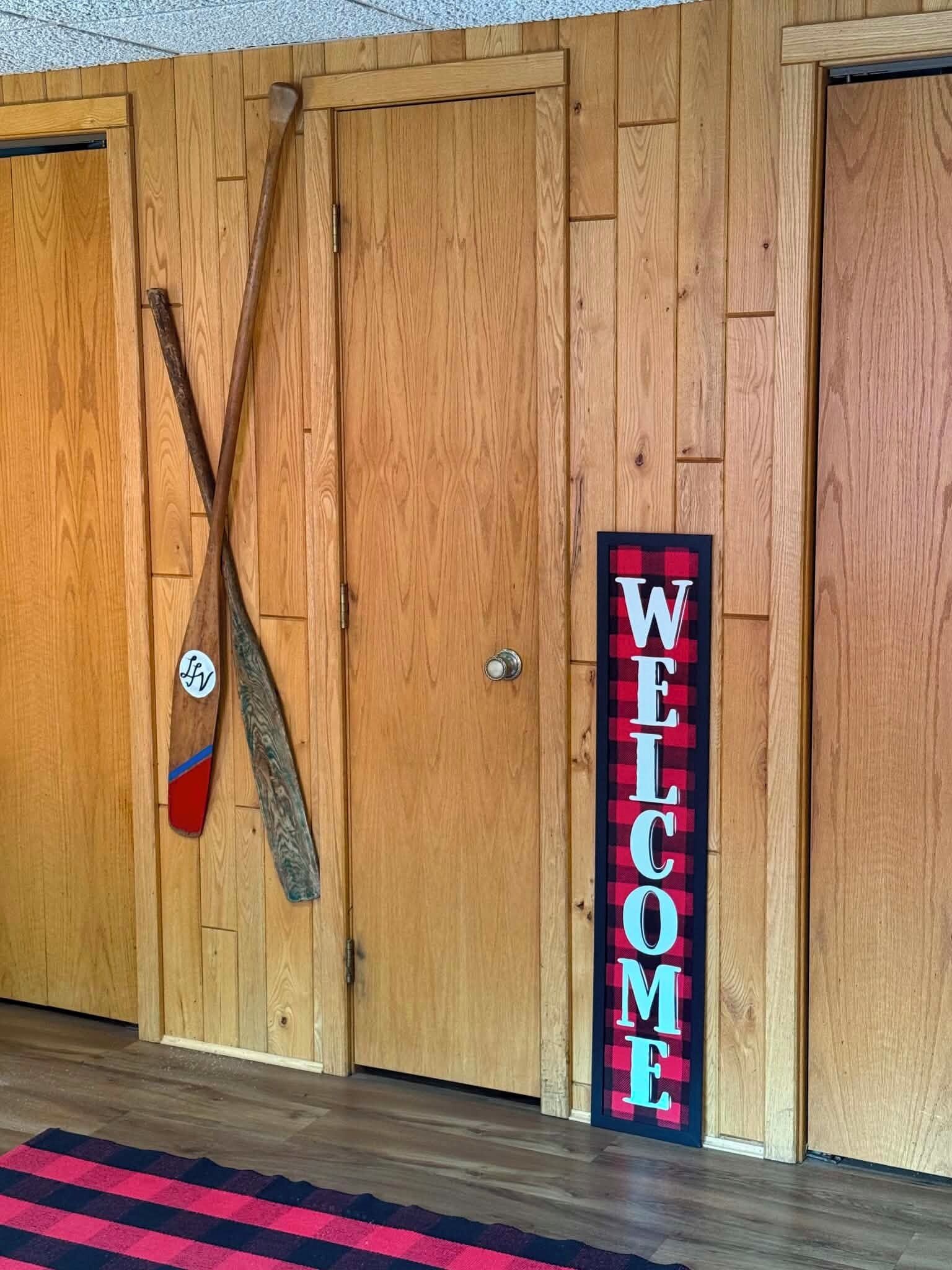 Une porte en bois, ornée d’une pancarte accueillante, et un bâton de marche en bois appuyé contre celle-ci.