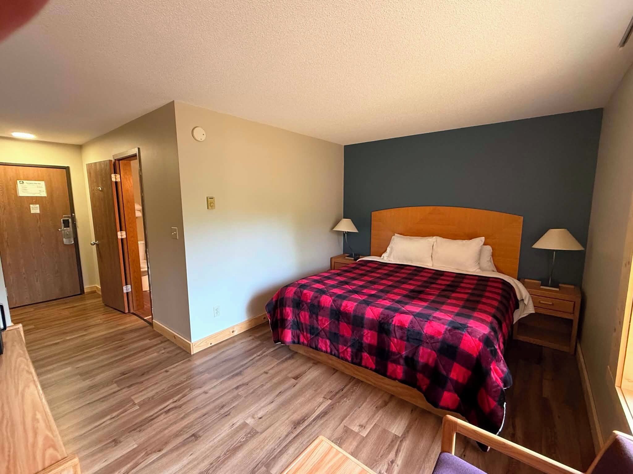 A hotel room with a wooden floor, a bed with a red and black checkered bedspread, two bedside lamps, and a nightstand.