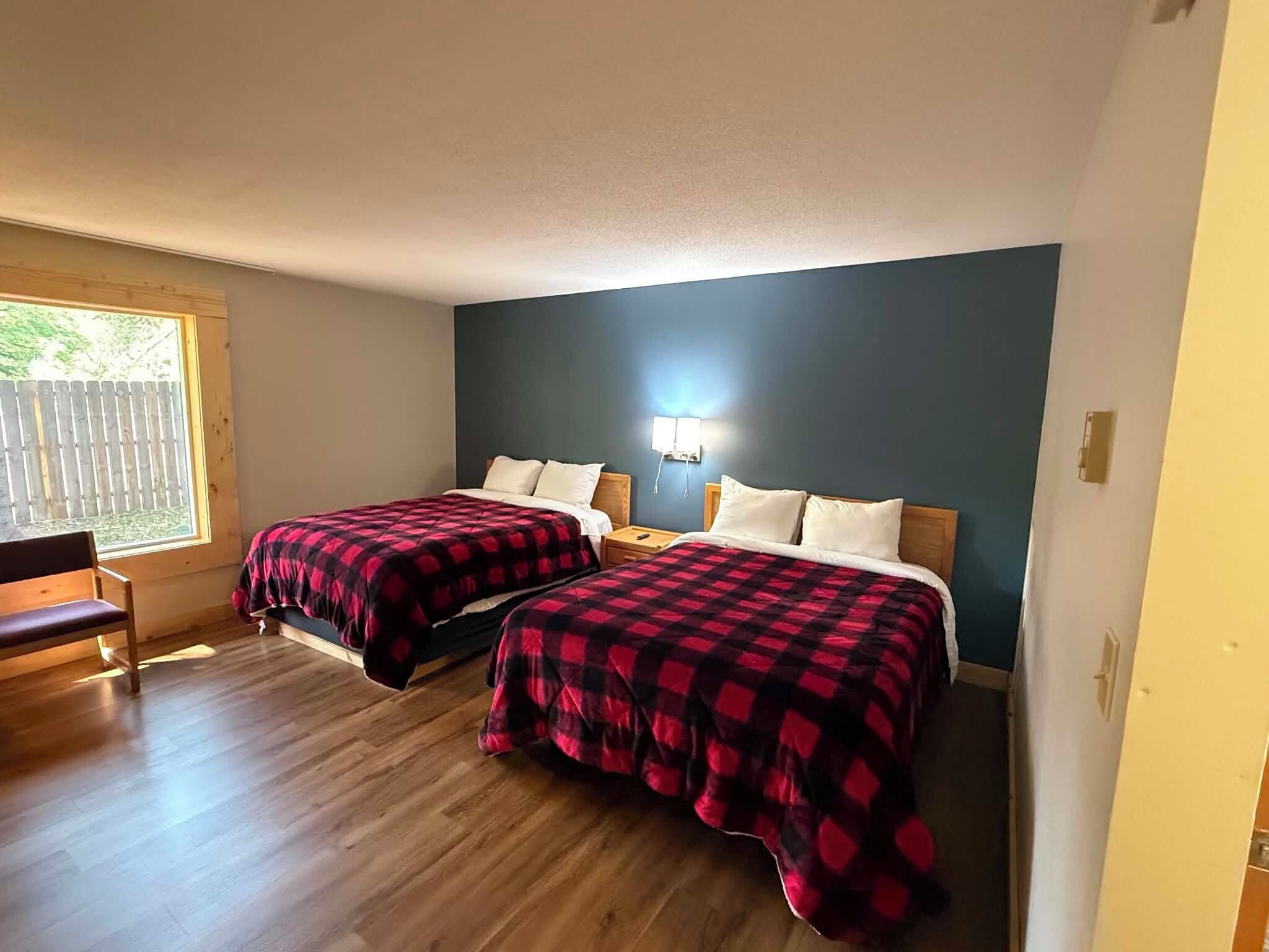 A room with two beds, wooden flooring, a bench, and a window with a view.