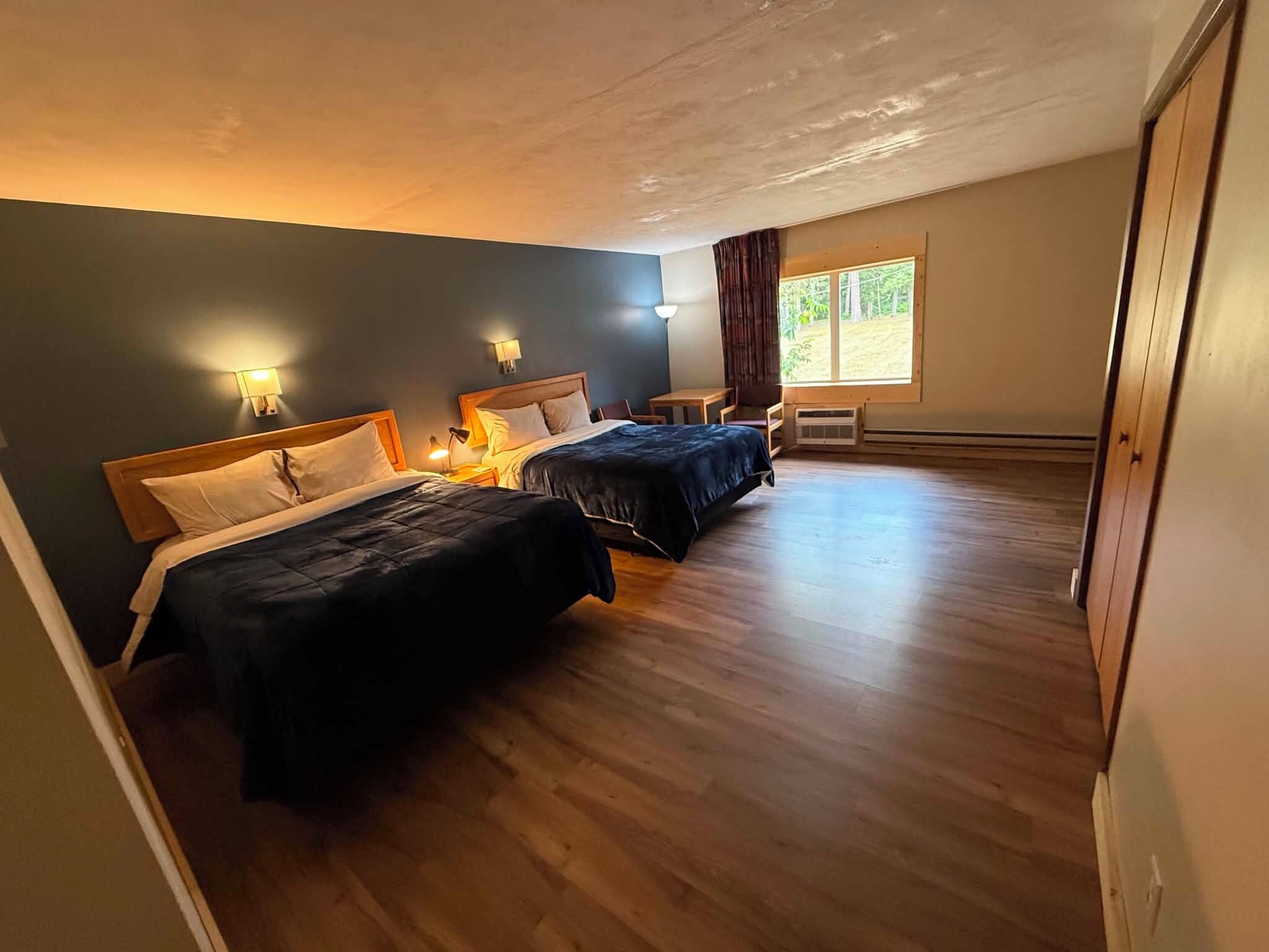A room with two beds, wooden flooring, and a window with curtains.