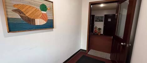 A hallway with a wooden duck painting, a dark mat at the entrance, and a door leading to a room with a plant and a small statue.