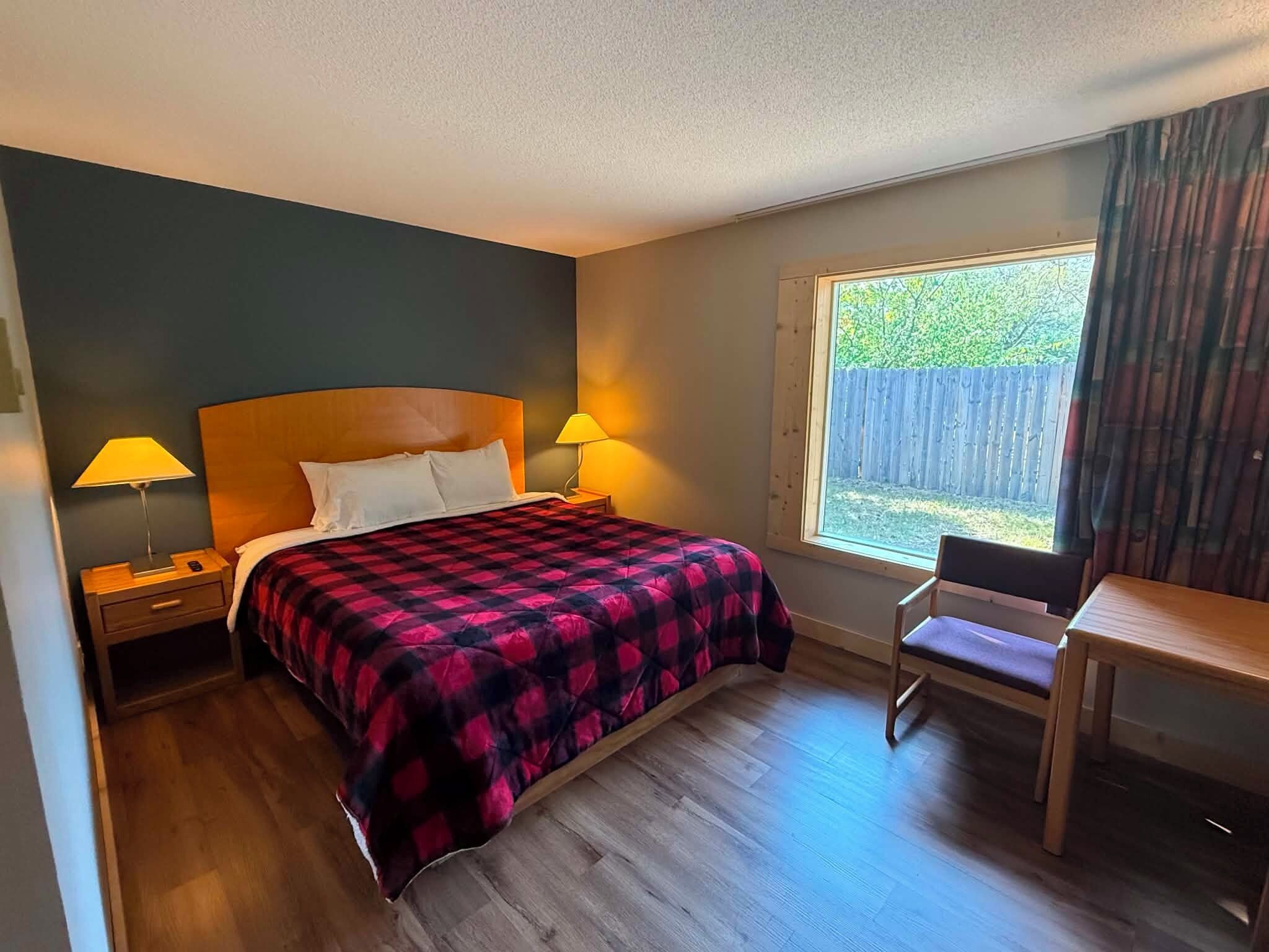 Un dormitorio con una cama, dos lámparas de noche, una mesita de noche de madera, una silla y una ventana con vistas a una valla y follaje.