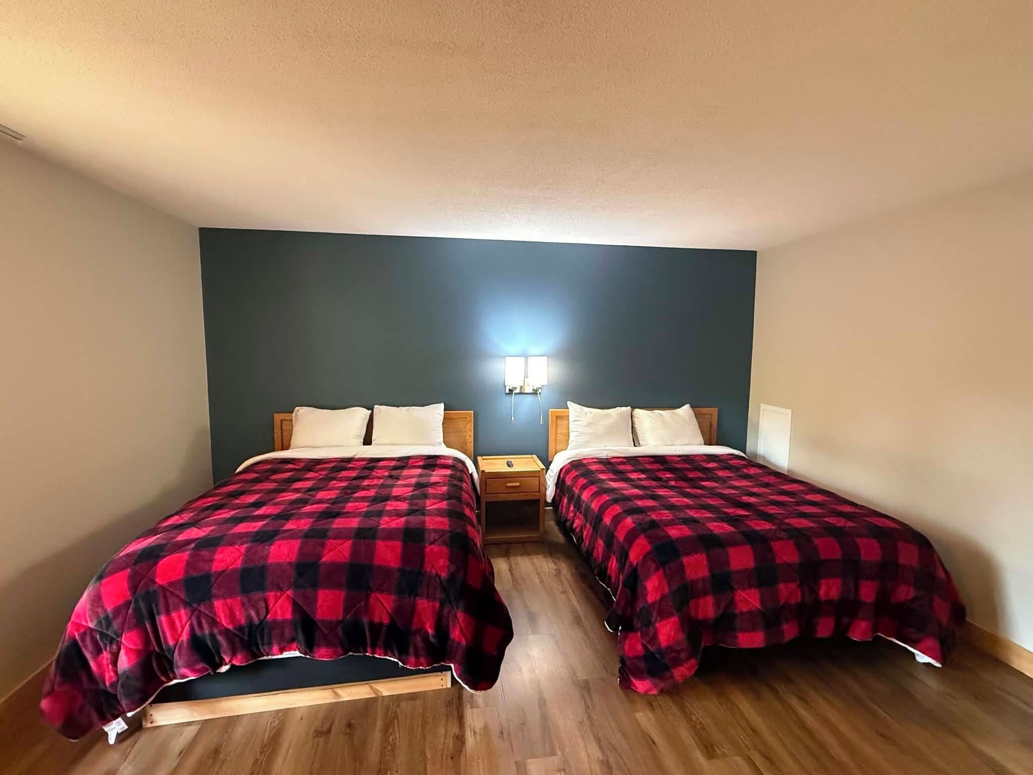 A room with two beds, each with a red and black checkered bedspread, wooden headboards, and a nightstand with a lamp between them.