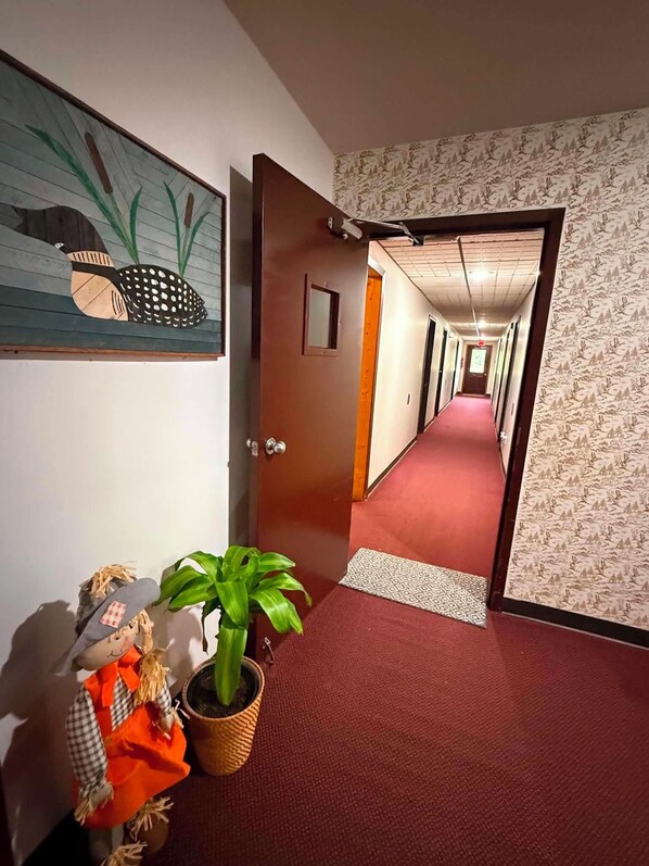 Pasillo de hotel con alfombra roja, una puerta de madera y un papel tapiz con motivos florales.