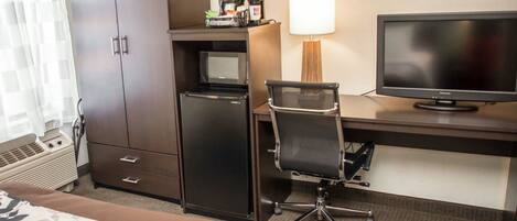 A hotel room with a bed, a desk with a computer and TV, a minibar, and a lamp.