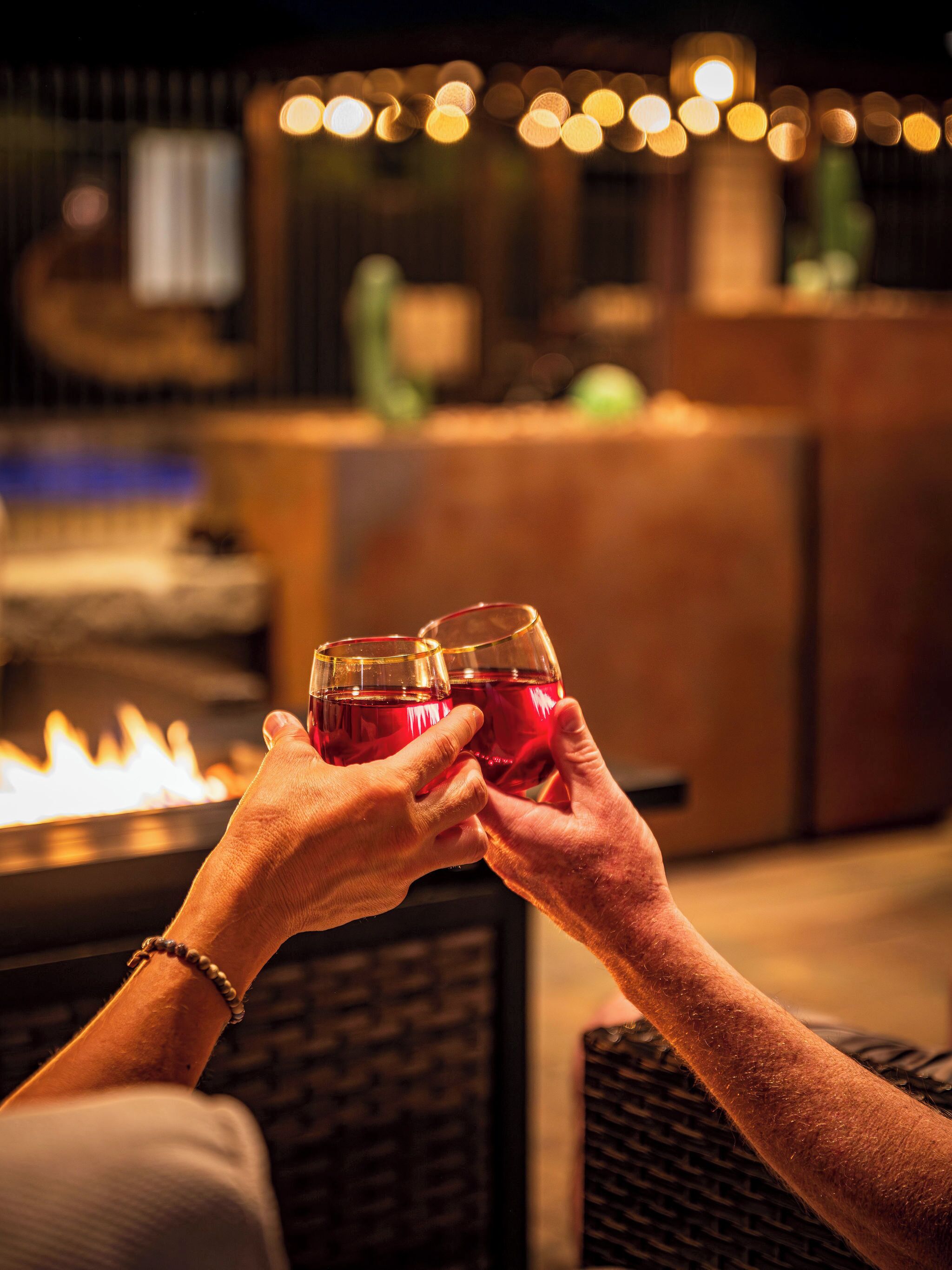 Two hands holding glasses of red liquid, a fire feature, and a bar area with lights in the background.
