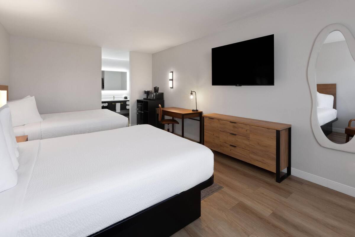 A modern hotel room with two beds, a desk, a flat-screen TV, a mirror, and a wooden dresser.