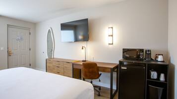 A hotel room with a bed, a wooden desk, a chair, a television mounted on the wall, a microwave, and a coffee maker.