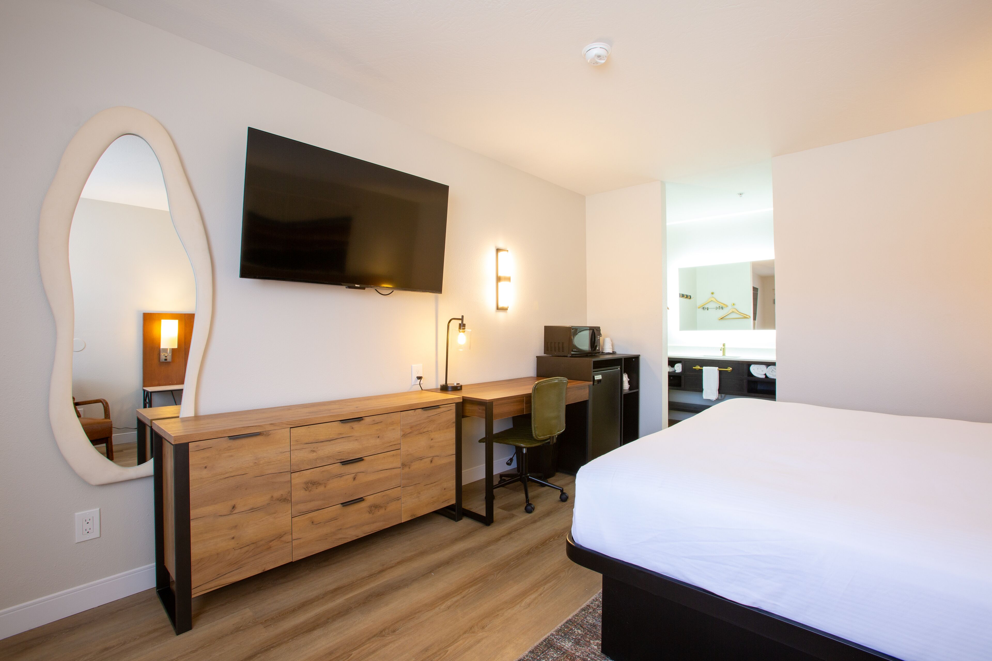 A modern hotel room with a wooden desk, a flat-screen TV, a large bed, and a mirror.