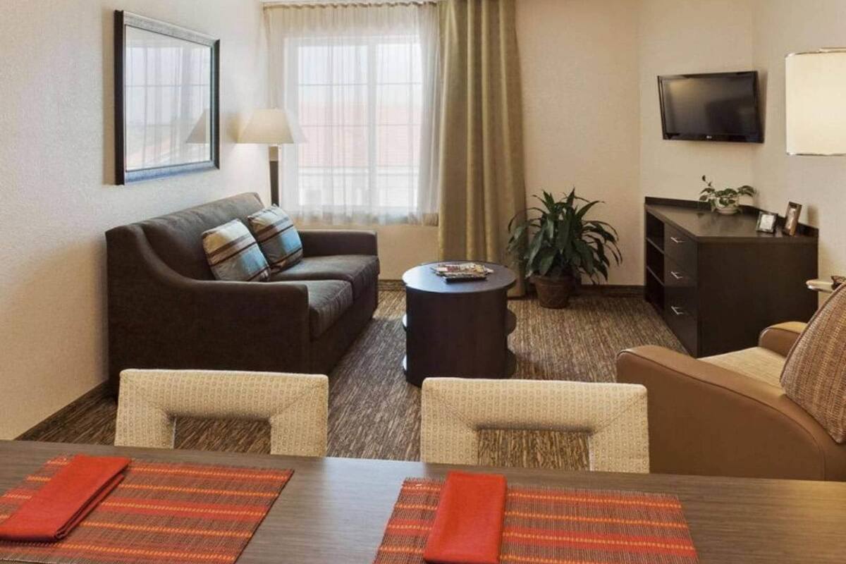 Habitación de hotel con sofá, sillones, mesa de comedor, televisor, lámpara, planta y ventana con cortinas.