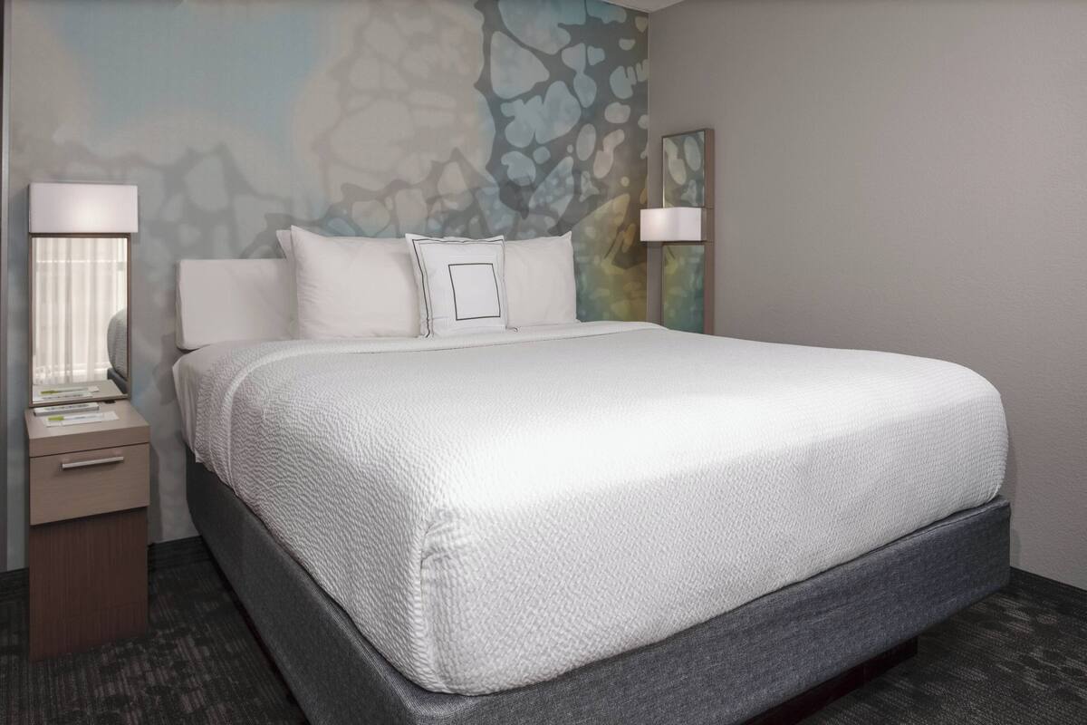 A neatly made bed with a white textured bedspread and pillows, a bedside table with a lamp, and a wall with a textured abstract design.