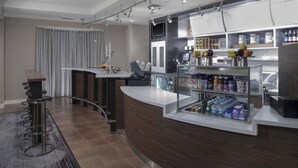 Un moderno vestíbulo de hotel con un mostrador de recepción curvo, un área de bar con taburetes, una vitrina de vidrio con bebidas y un panel de menú sobre el mostrador.