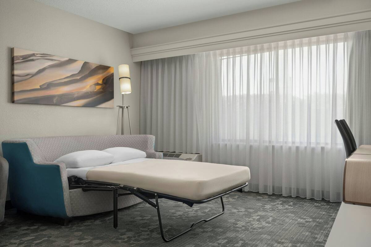 A hotel room with a bed, a desk, a chair, a lamp, a large window with curtains, and a painting on the wall.