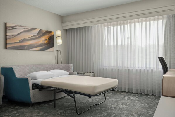 A hotel room with a bed, a desk, a chair, a lamp, a large window with curtains, and a painting on the wall.