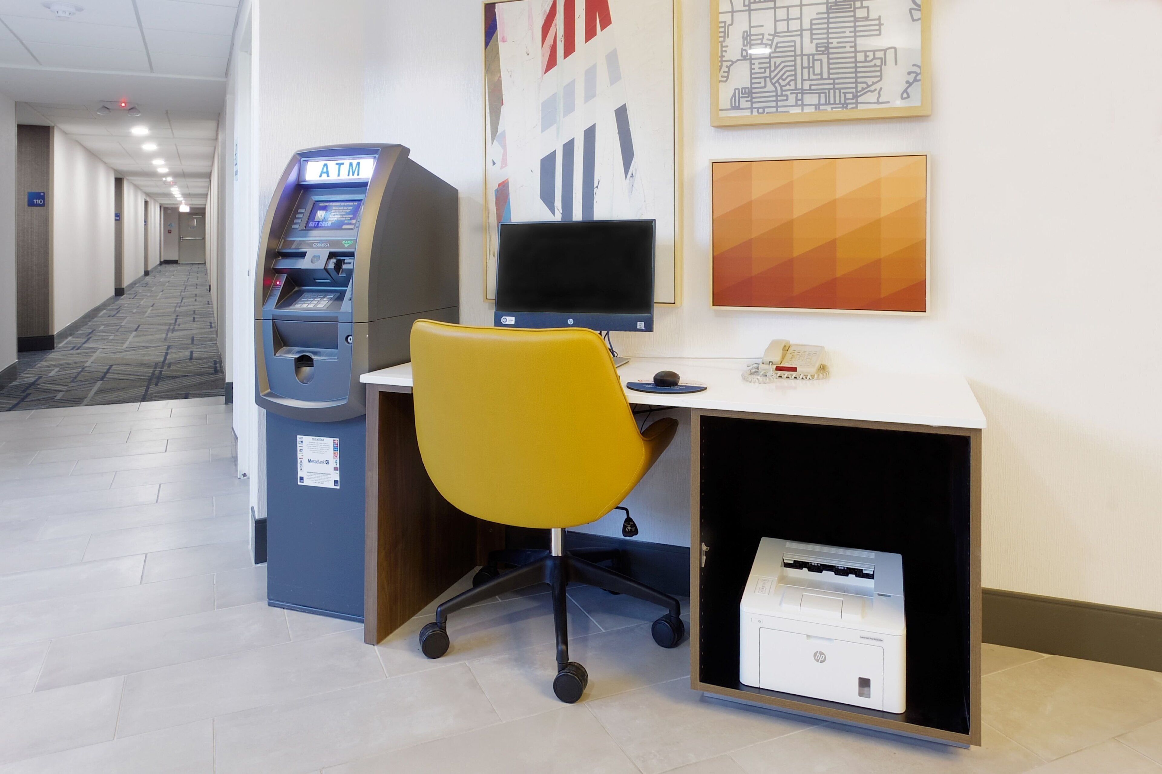 A hotel lobby with an ATM, a desk with a computer, a yellow office chair, and a printer.