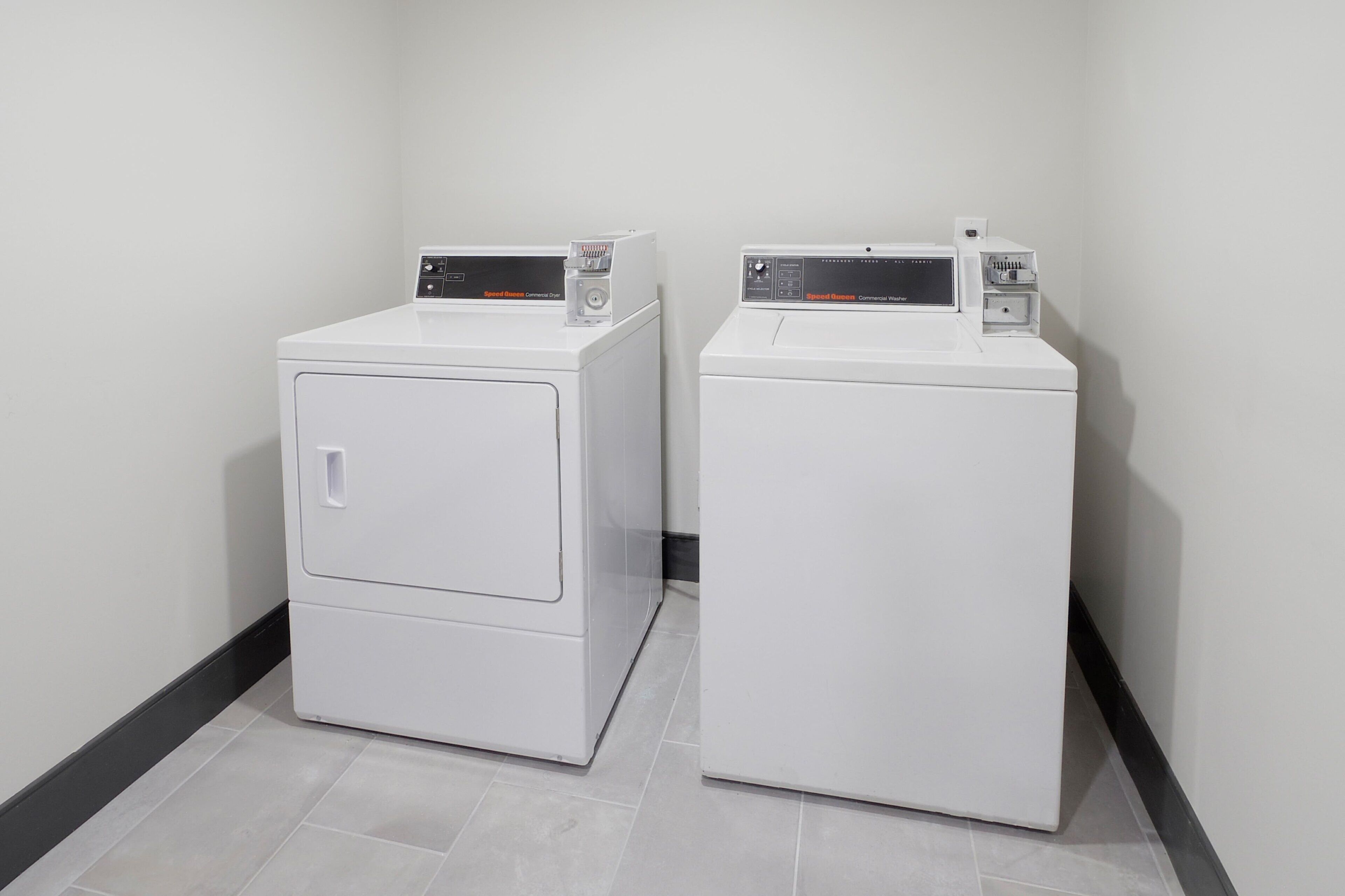 Two white front-loading washing machines in a corner of a room with light-colored tiled flooring and a black baseboard.