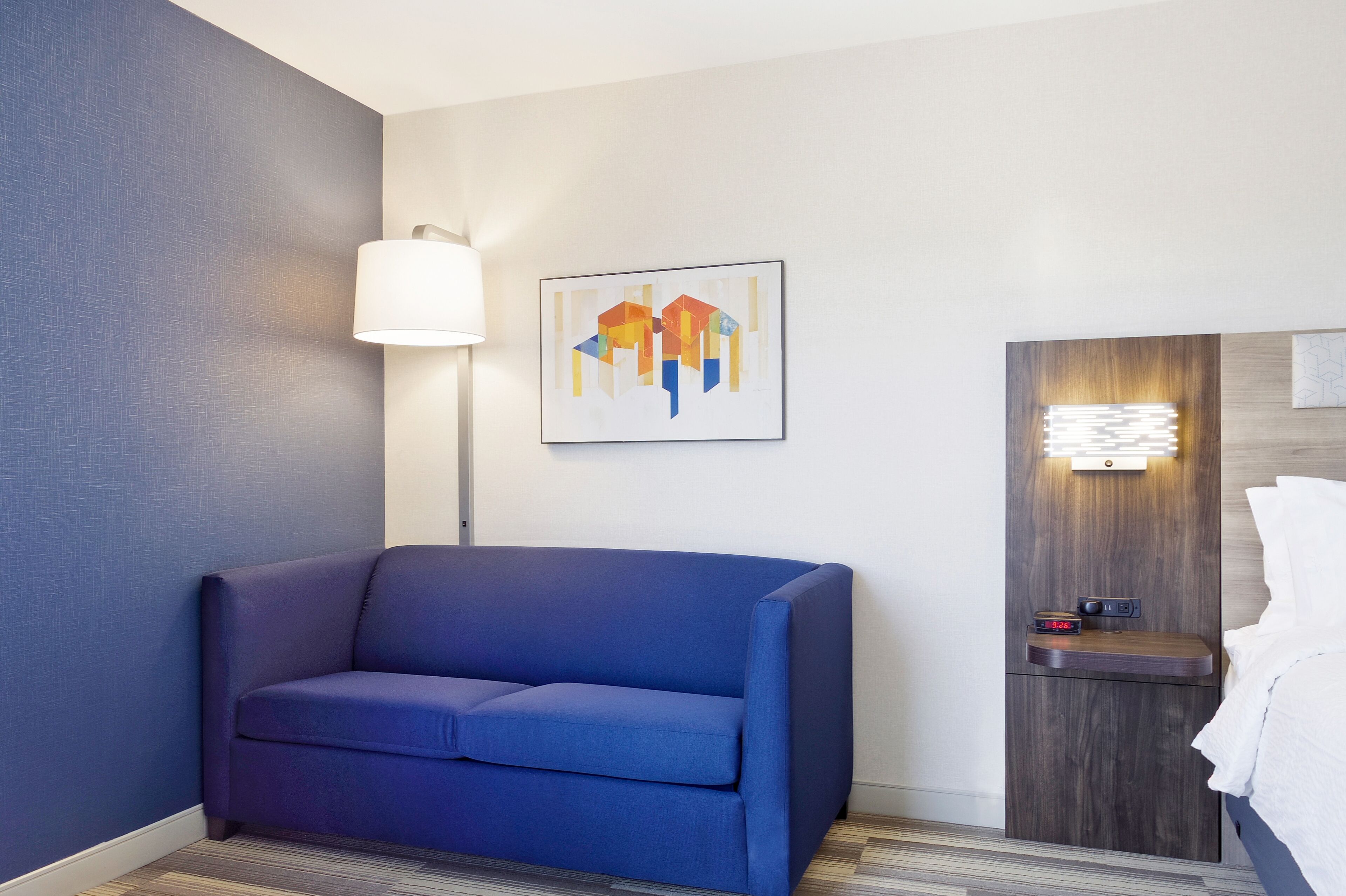 Une chambre d’hôtel moderne avec un canapé bleu, une lampe fixée au mur, une œuvre d’art abstraite encadrée, une table de chevet avec une lampe et une tête de lit en bois.