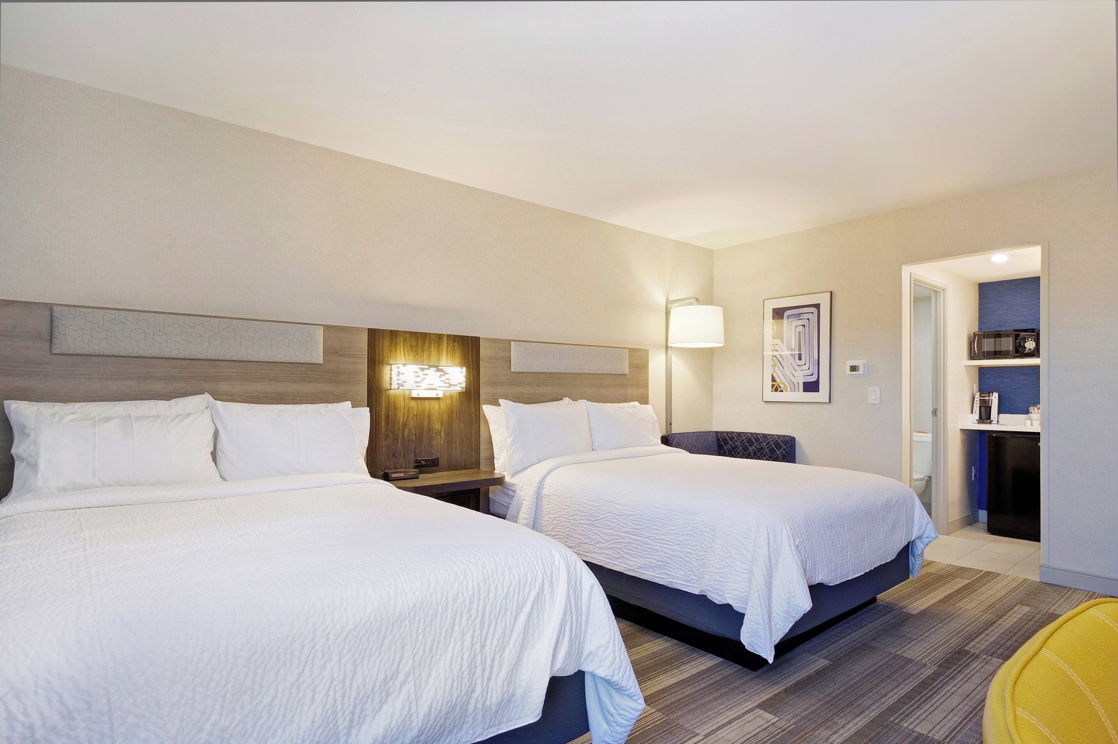Habitación de hotel con dos camas, una mesita de noche, una lámpara de pared, una pequeña mesa y un microondas en una esquina.