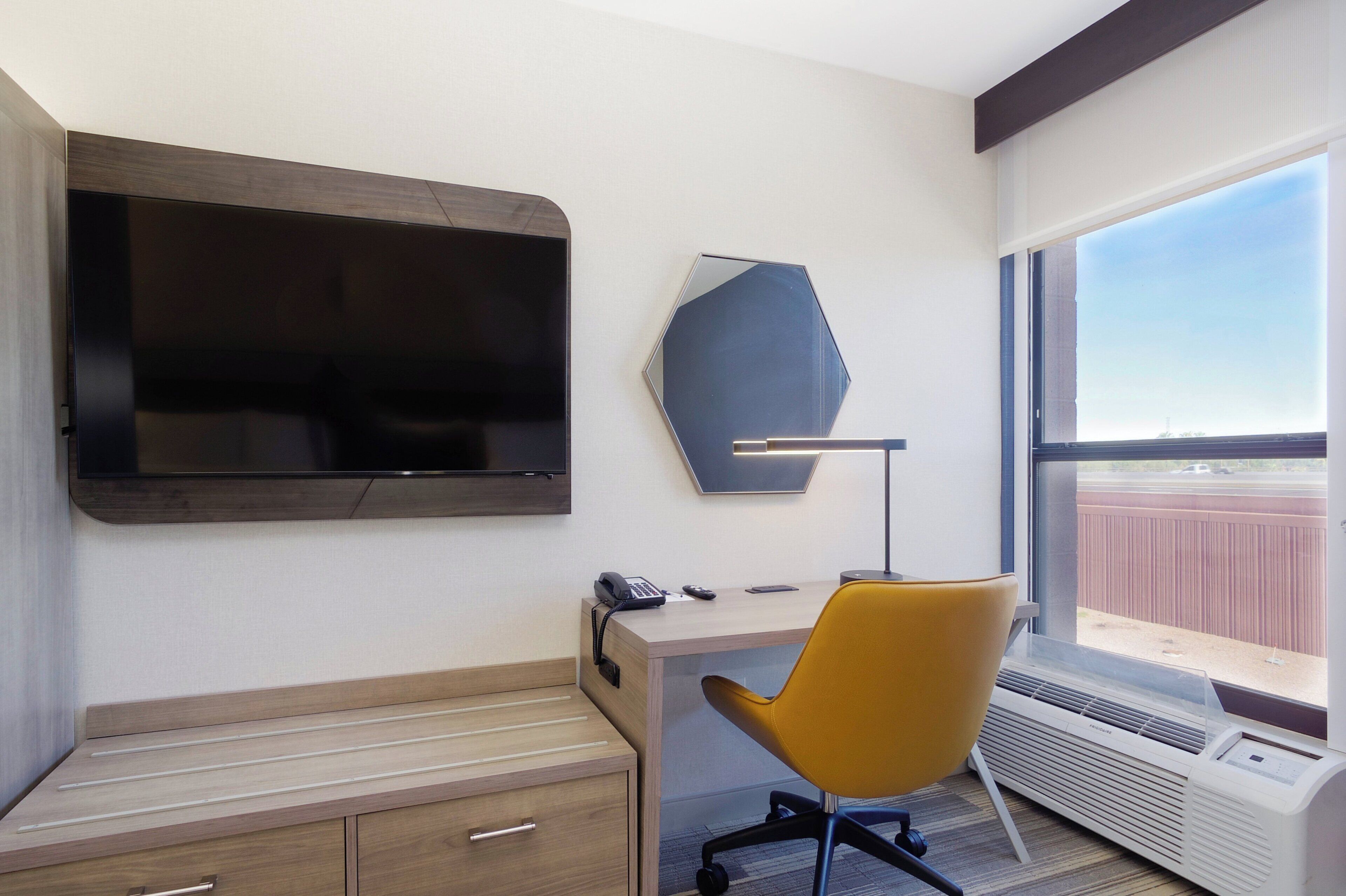 A hotel room with a large flat-screen TV, a desk with a chair, a telephone, and a window with a view.
