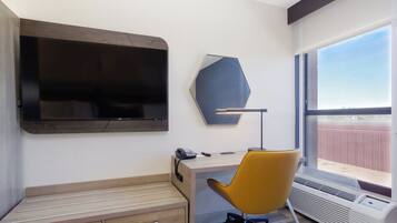 A hotel room with a large flat-screen TV, a desk with a chair, a telephone, and a window with a view.