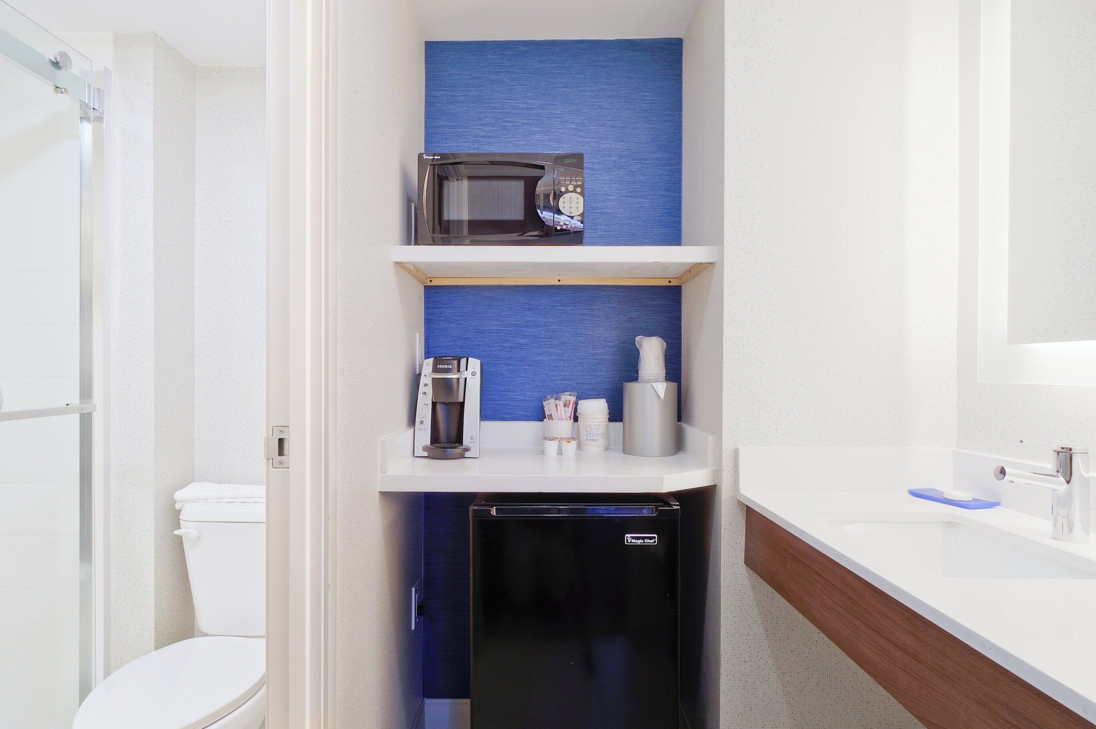 A compact bathroom with a toilet, a coffee machine, a microwave, and a small refrigerator.