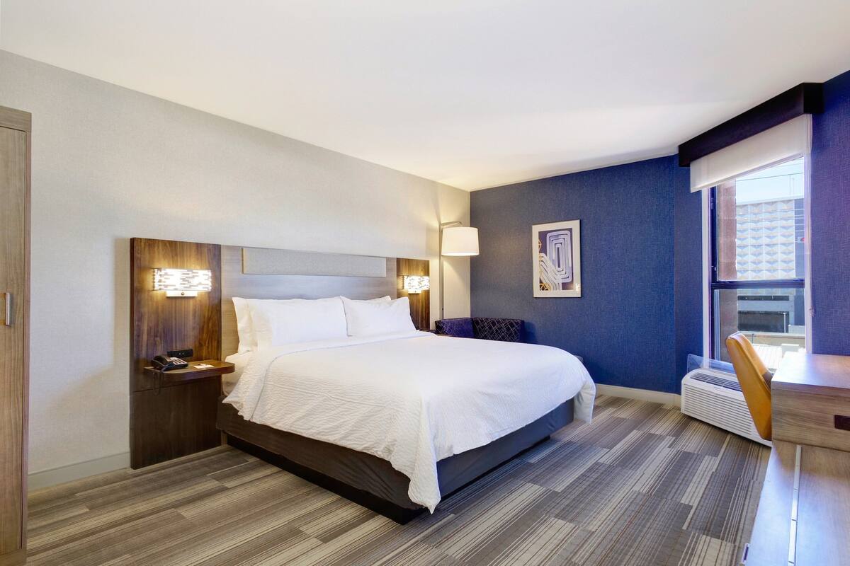 Une chambre d’hôtel avec un grand lit, des tables de chevet, un bureau et une fenêtre avec des stores.
