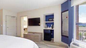 A hotel room with a bed, a TV mounted on the wall, a small kitchenette with a microwave, and a window with a view.