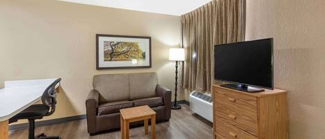 Quarto de hotel com uma escrivaninha, um sofá de couro marrom, uma pequena mesa de madeira, uma televisão sobre suporte de madeira, um quadro pendurado na parede e um abajur.