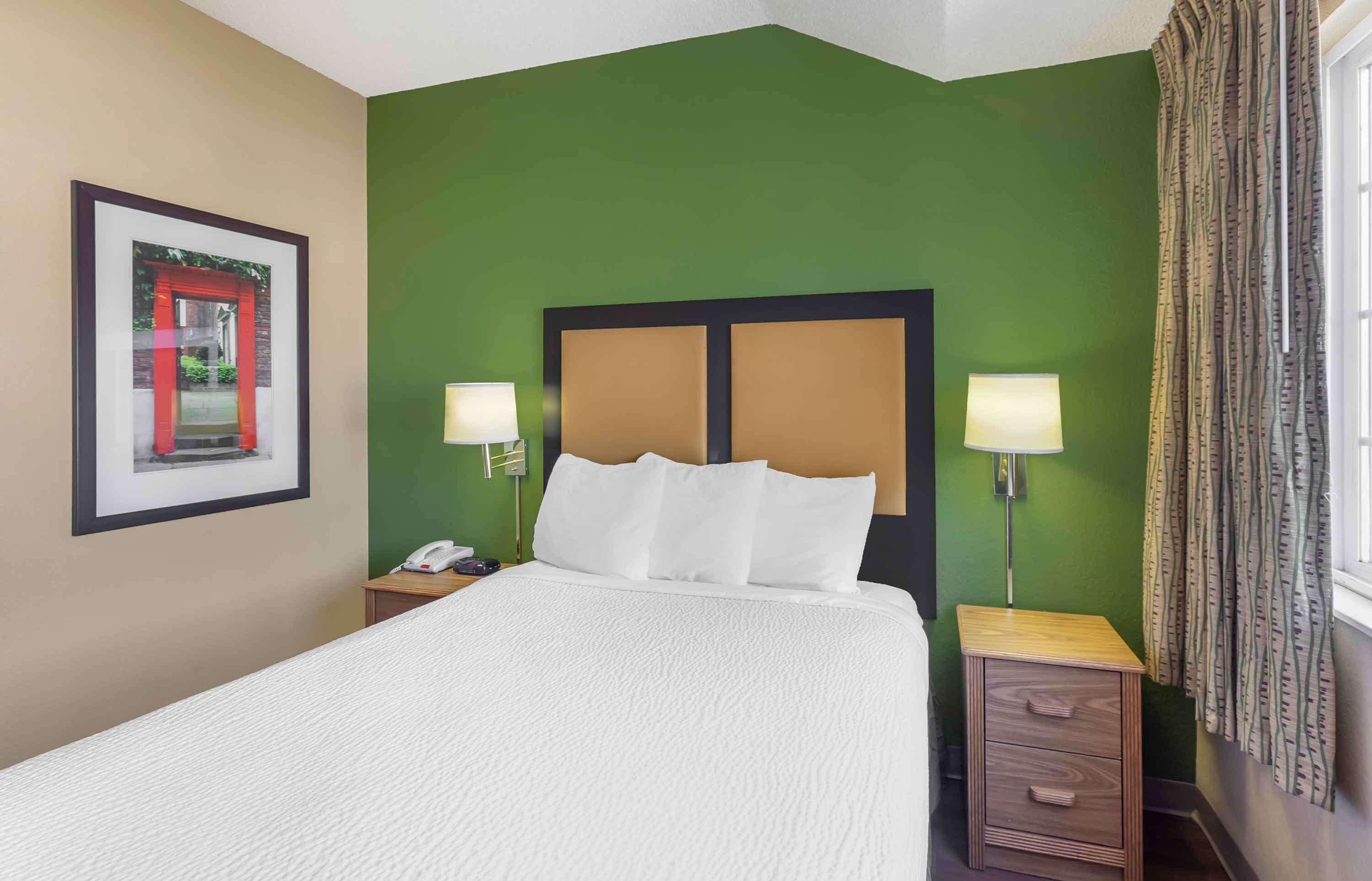 A hotel room with a large bed, green walls, two bedside lamps, a framed picture on the wall, and a window with curtains.