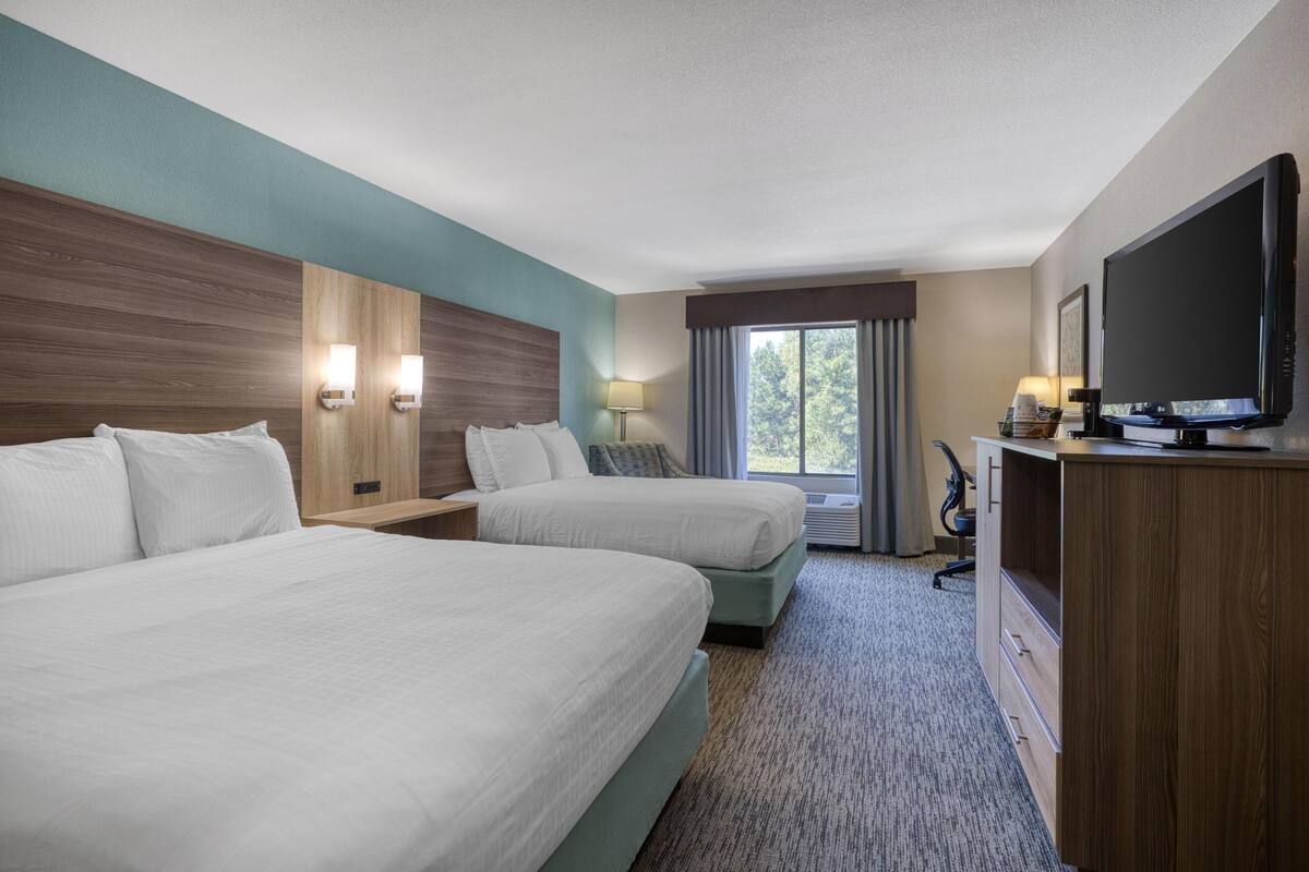 A hotel room with two beds, a TV, a desk, and a window with a view of trees.