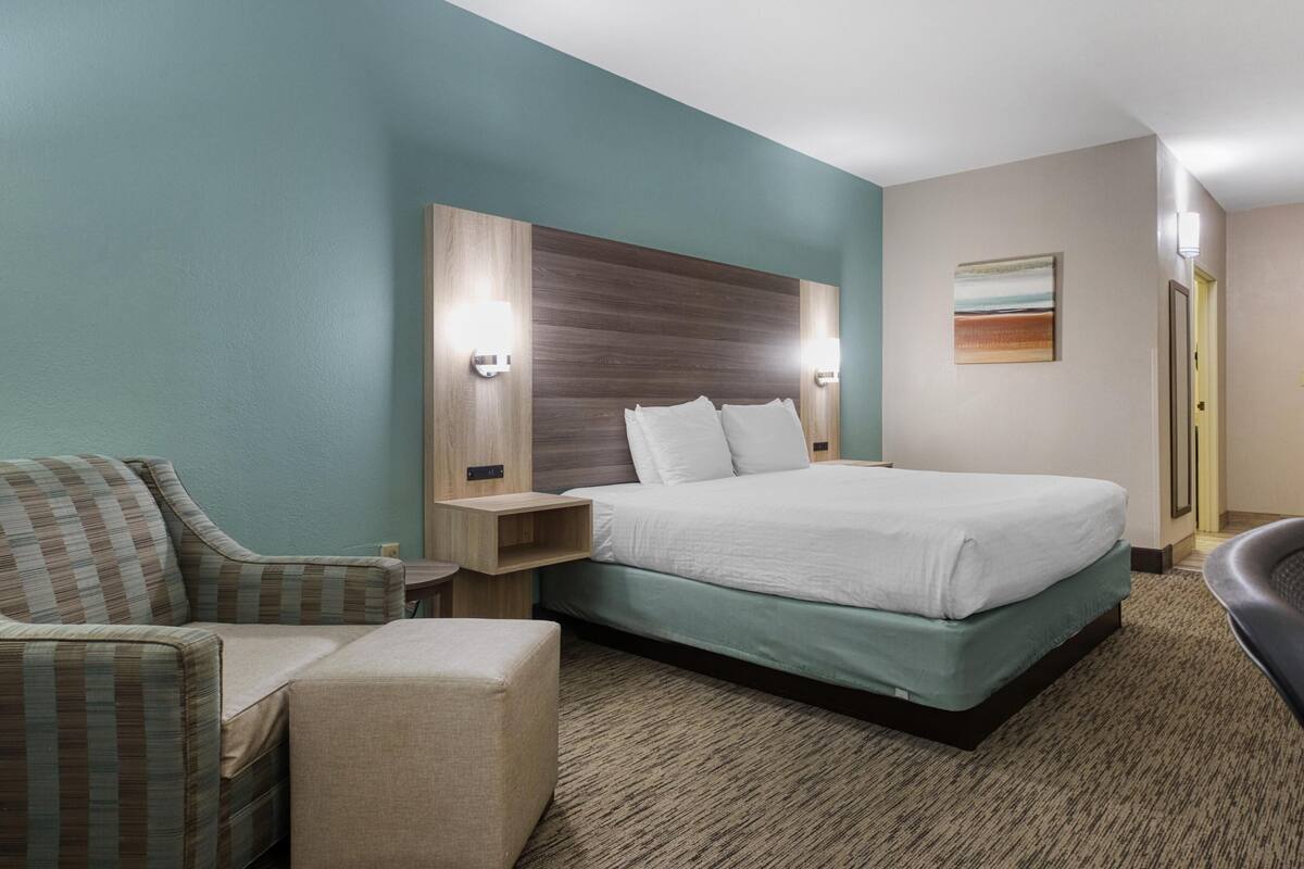 A hotel room with a bed, a chair, a nightstand, and a wall-mounted light fixture.