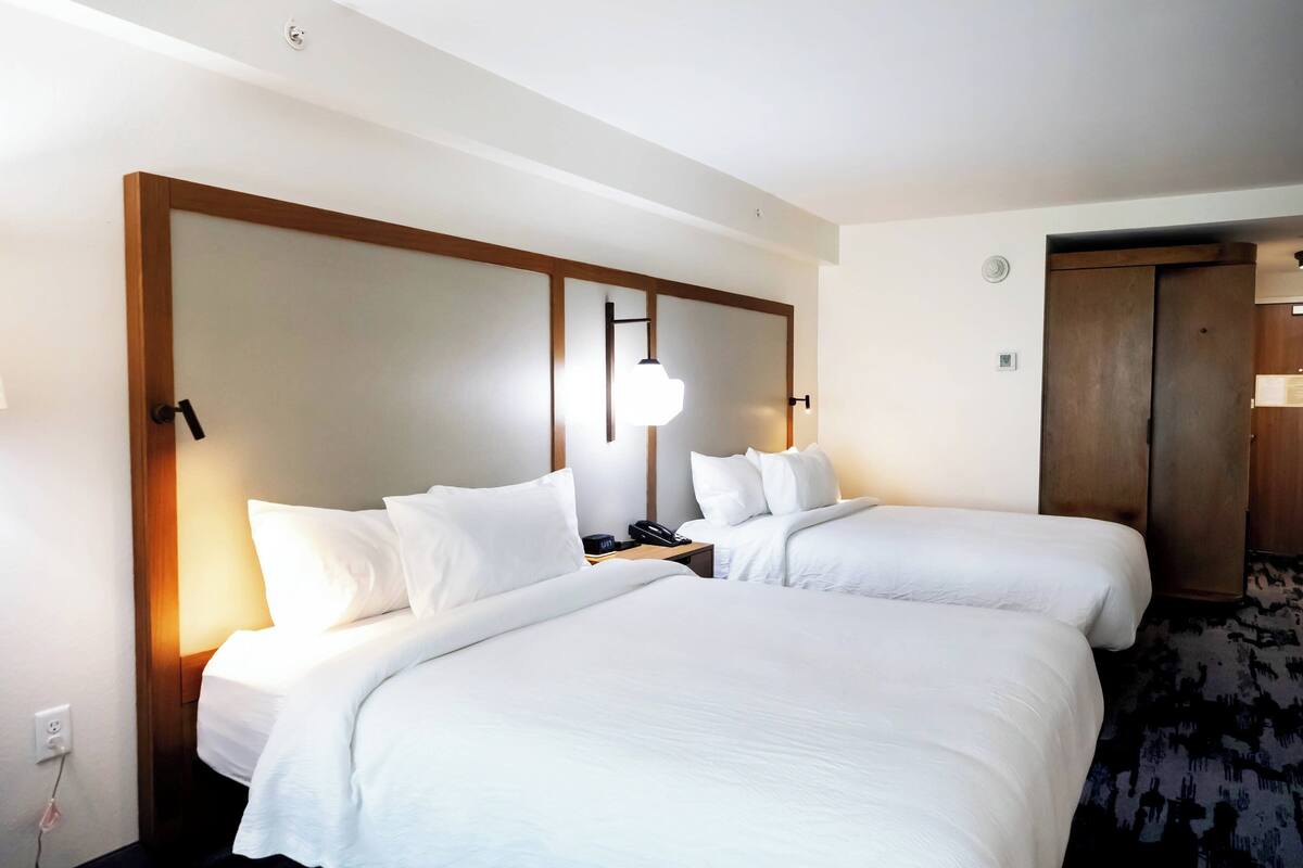 A hotel room with two beds, a wooden headboard, a bedside table with a lamp, and a door to the right.