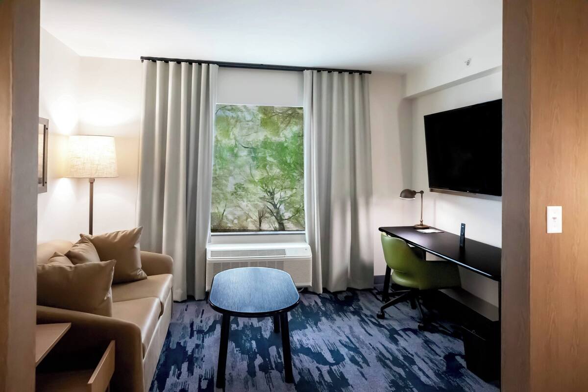 A hotel room with a sofa, a small table, a chair, a desk, a television, and a window with curtains.