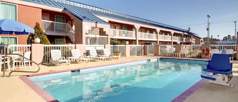 Un hotel con piscina, mobiliario de exterior y un edificio de dos pisos con balcones.