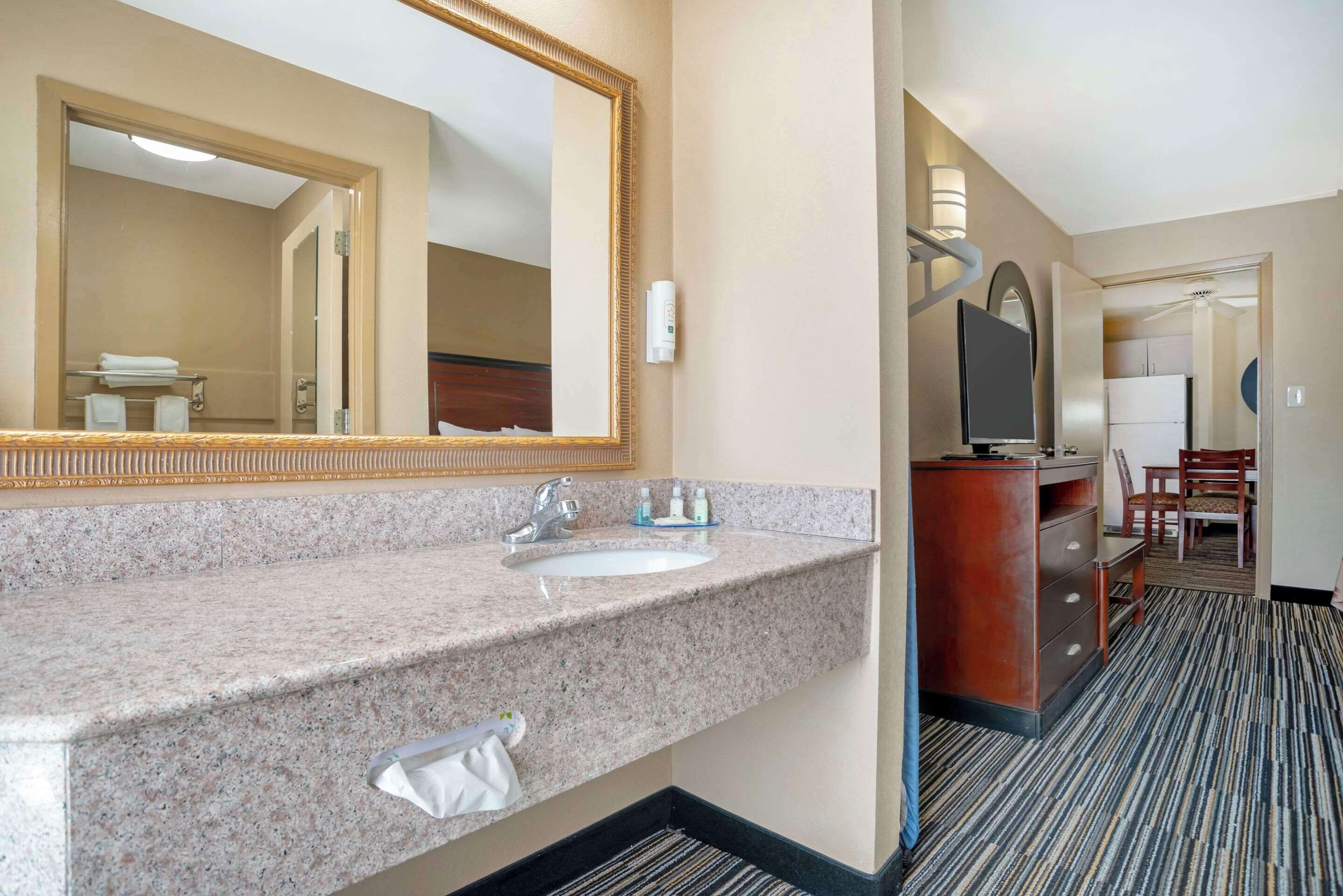 Un baño de hotel con lavamanos de encimera de granito, un espejo grande y un tocador con espejo. Se ve un televisor sobre un mueble de madera y una silla al fondo.