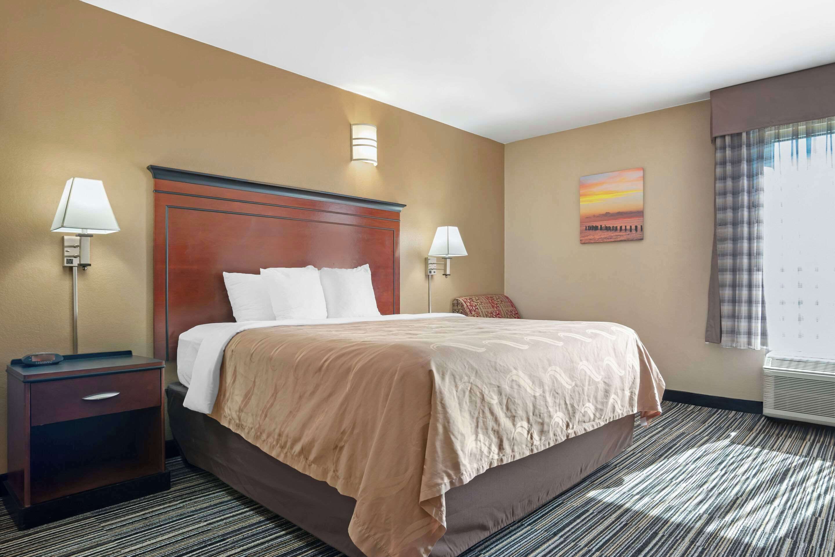 A hotel room with a large bed, two bedside lamps, a nightstand, a painting on the wall, and a window with curtains.