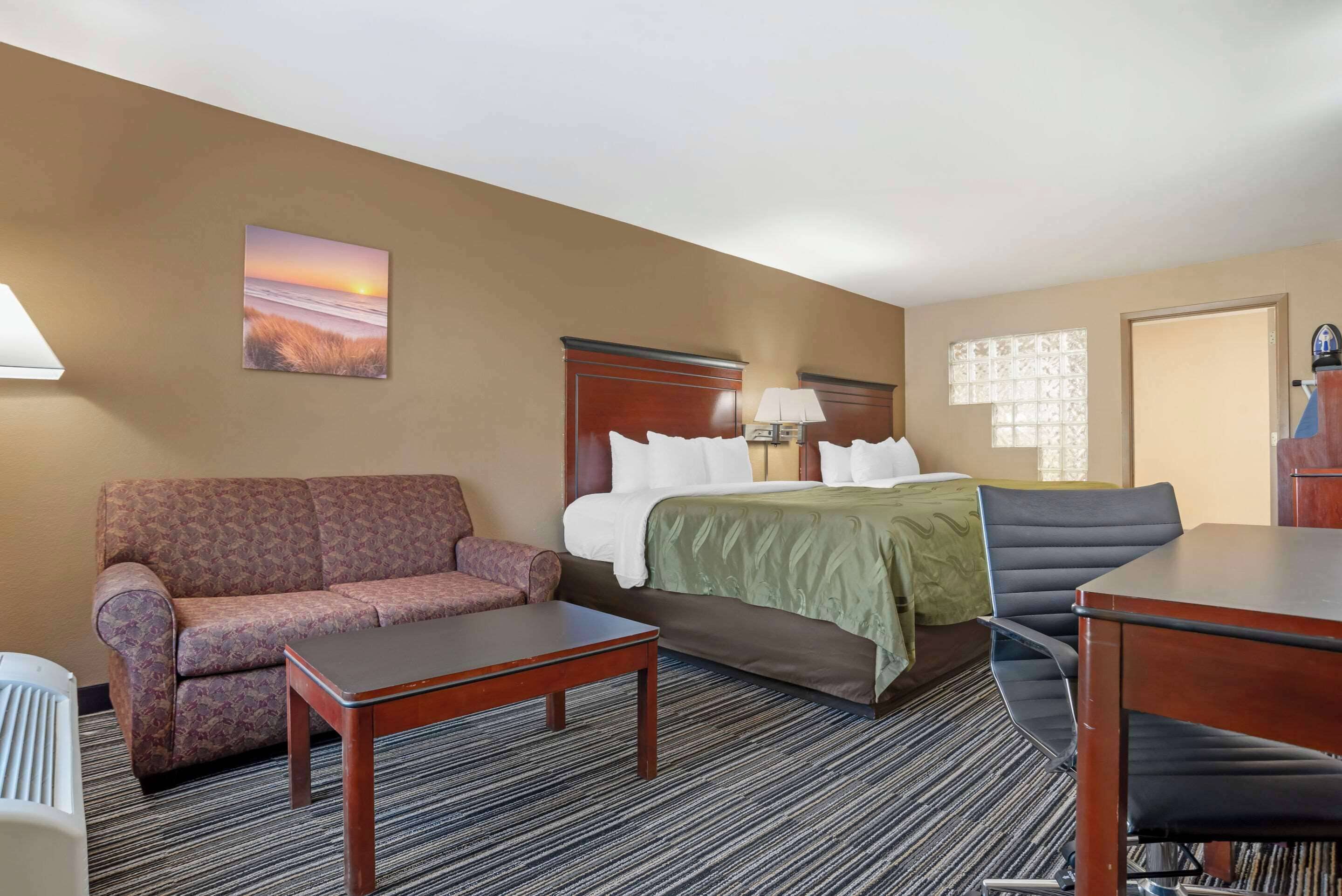 A hotel room with a bed, a sofa, a desk, and a chair. There is a painting on the wall and a window with blinds.