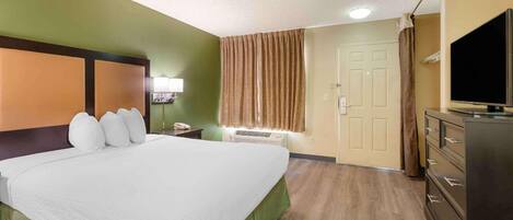 A hotel room with a bed, a TV, a dresser, a window with curtains, and an air conditioning unit.