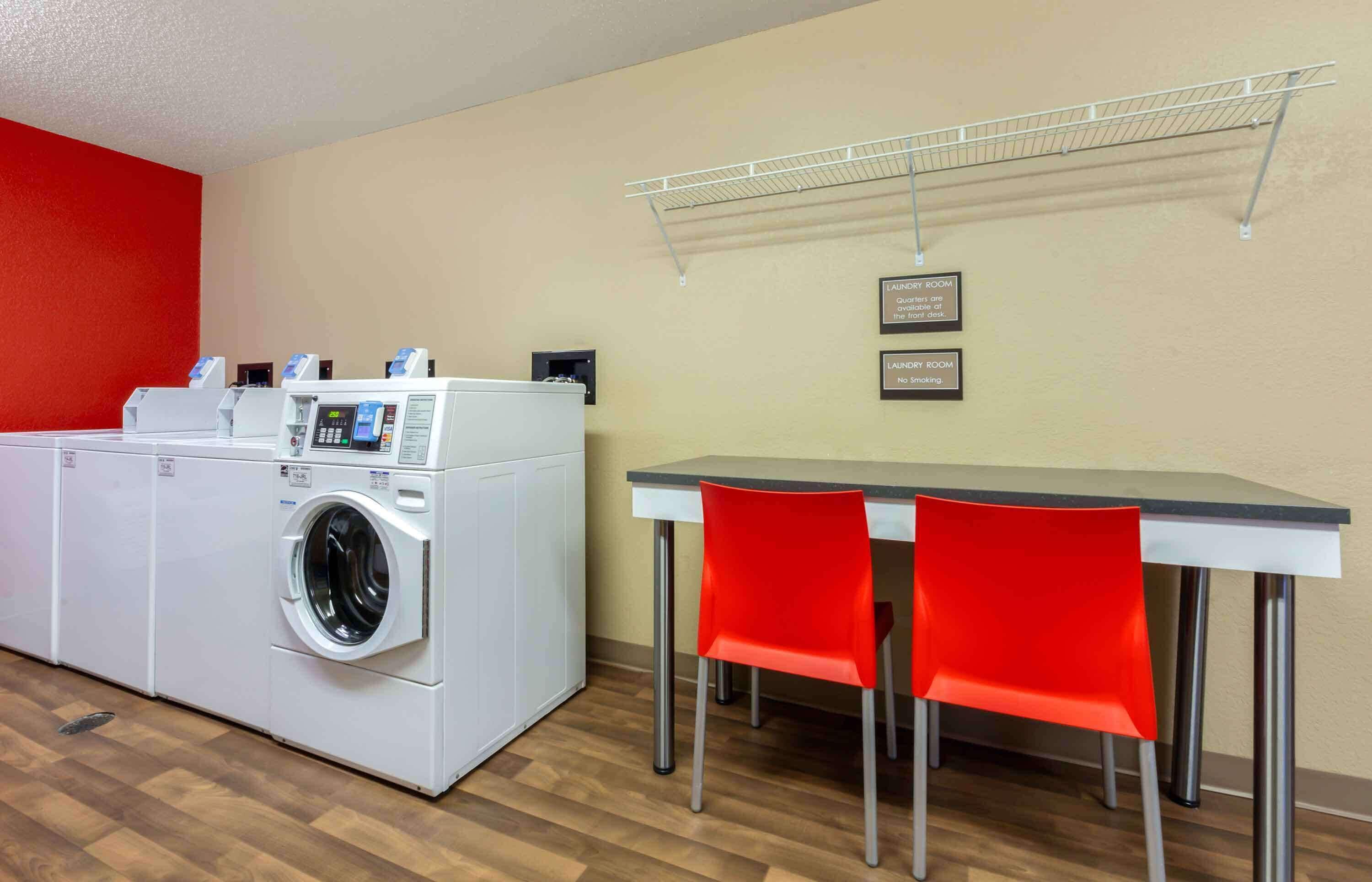 A laundry room with a washing machine and dryer, a table with two red chairs, and a metal drying rack on the wall.