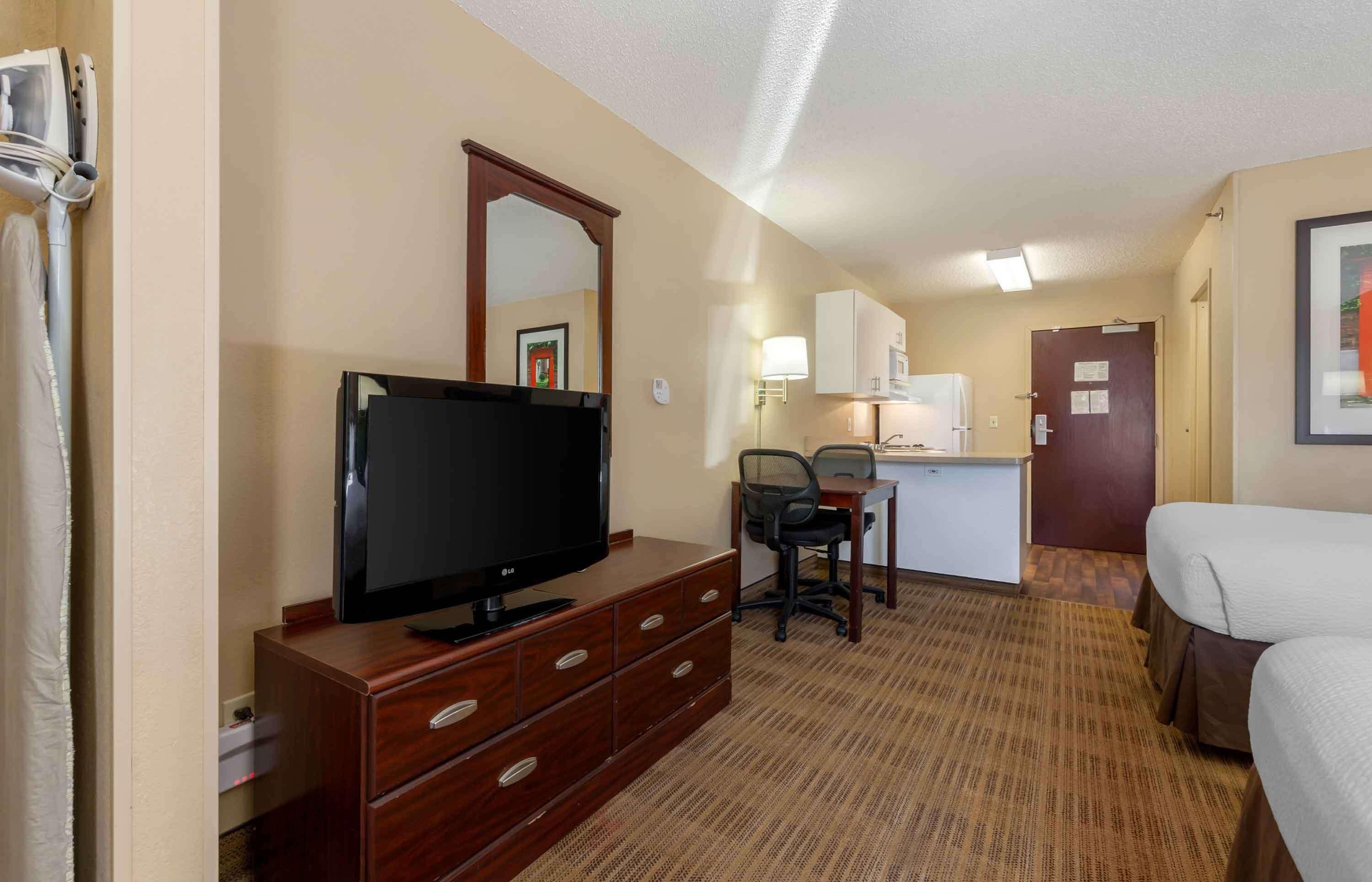 A hotel room with a flat-screen TV, wooden dresser, desk, chair, kitchenette, and a bed.