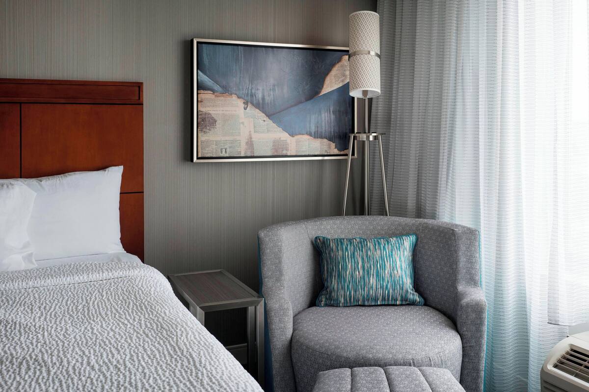 A hotel room with a bed, a gray armchair with a patterned pillow, a side table, a floor lamp, and a painting on the wall.