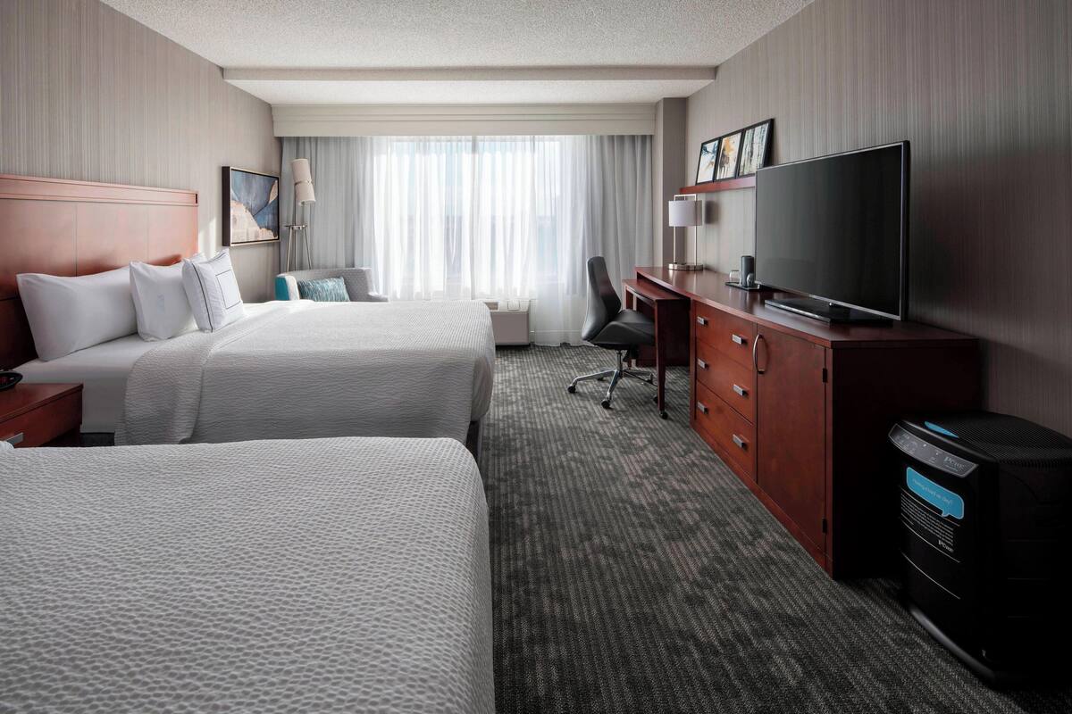 A hotel room with two beds, a desk, a television, and a large window with curtains.