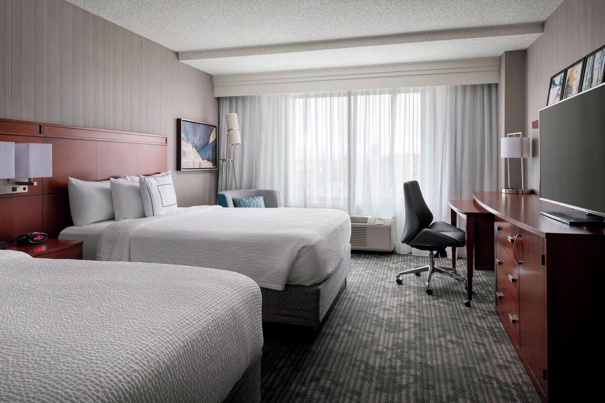A hotel room with two beds, a desk, a TV, and a window with curtains.