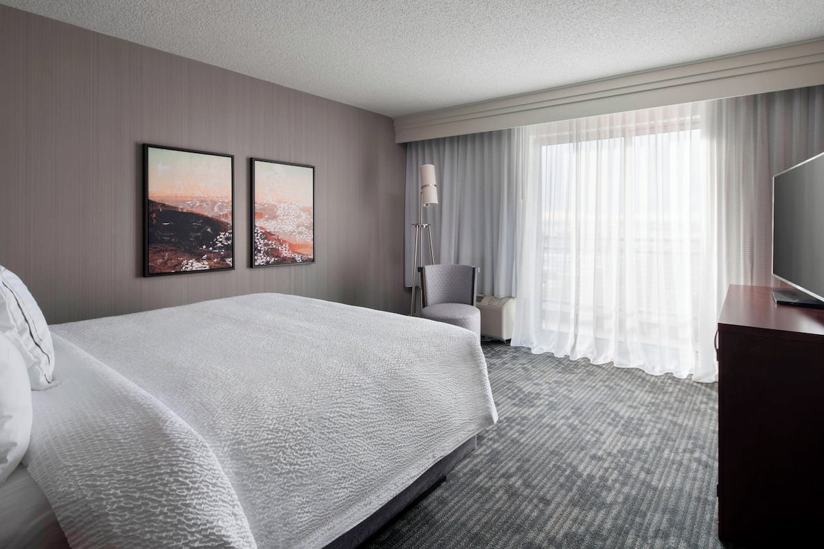 A hotel room with a large bed, two framed pictures on the wall, a chair, a desk with a flat-screen TV, and a large window with sheer curtains.