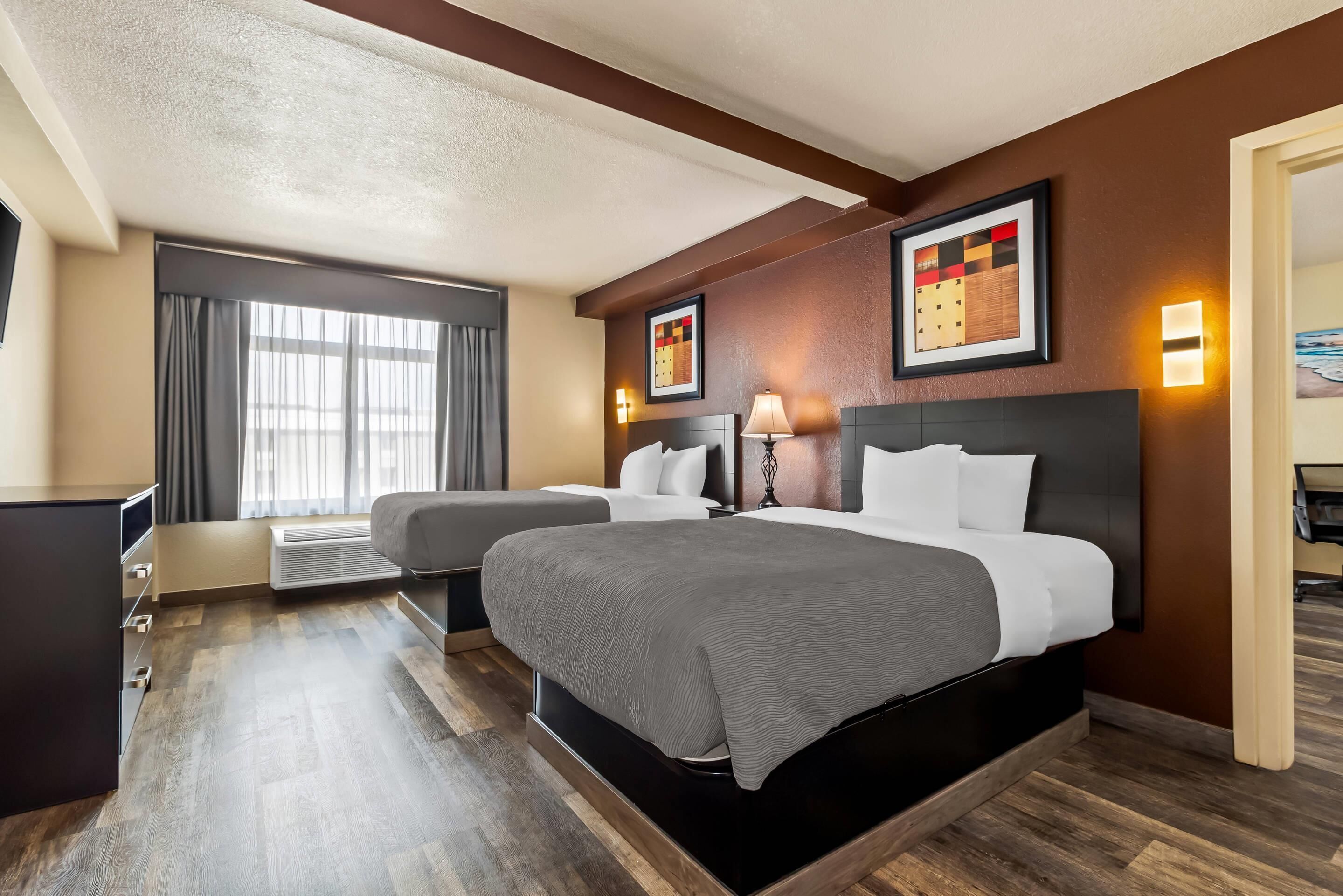 A hotel room with two beds, a desk, a chair, a window with curtains, and wall-mounted artwork.