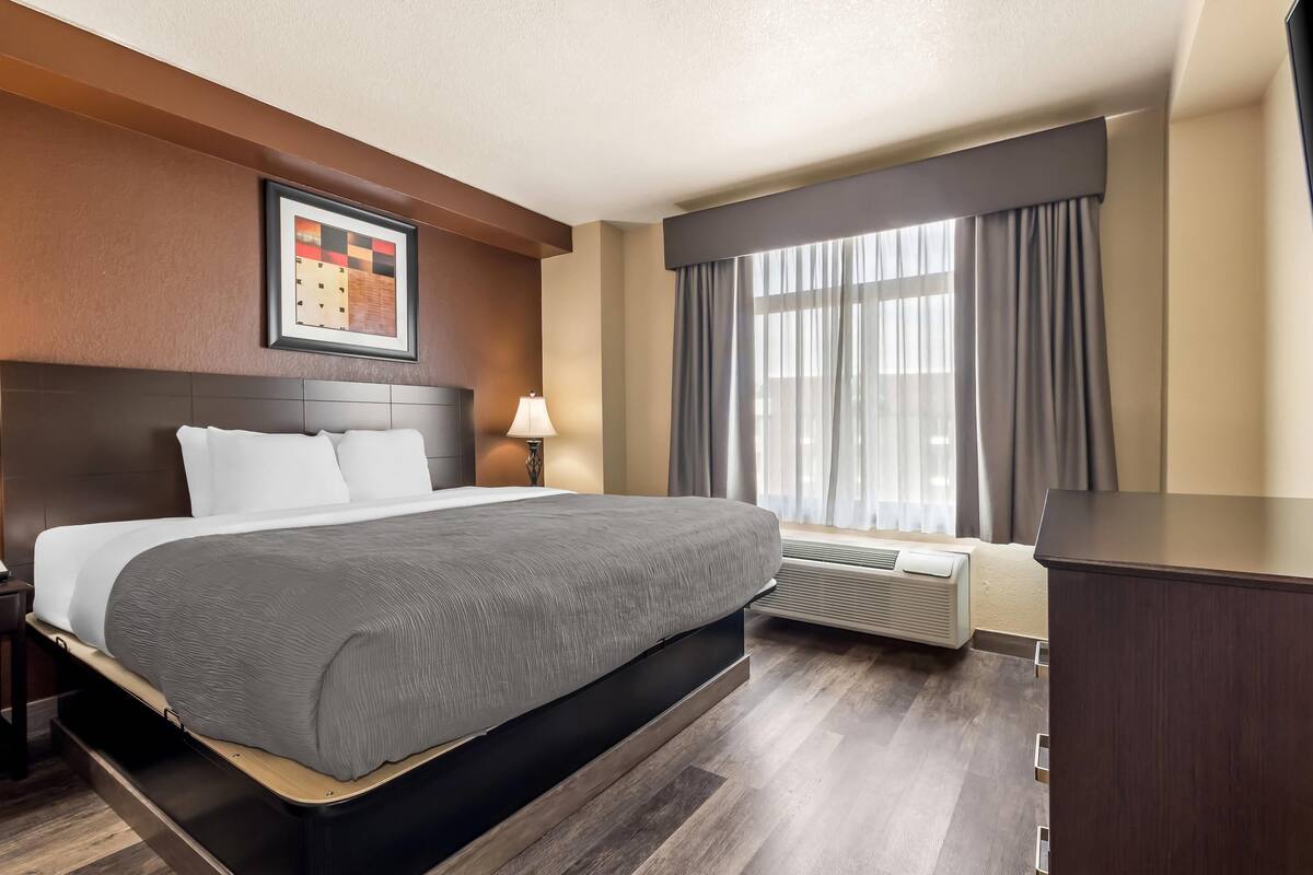 A hotel room with a bed, a nightstand, a lamp, a window with curtains, and a wall-mounted air conditioner.